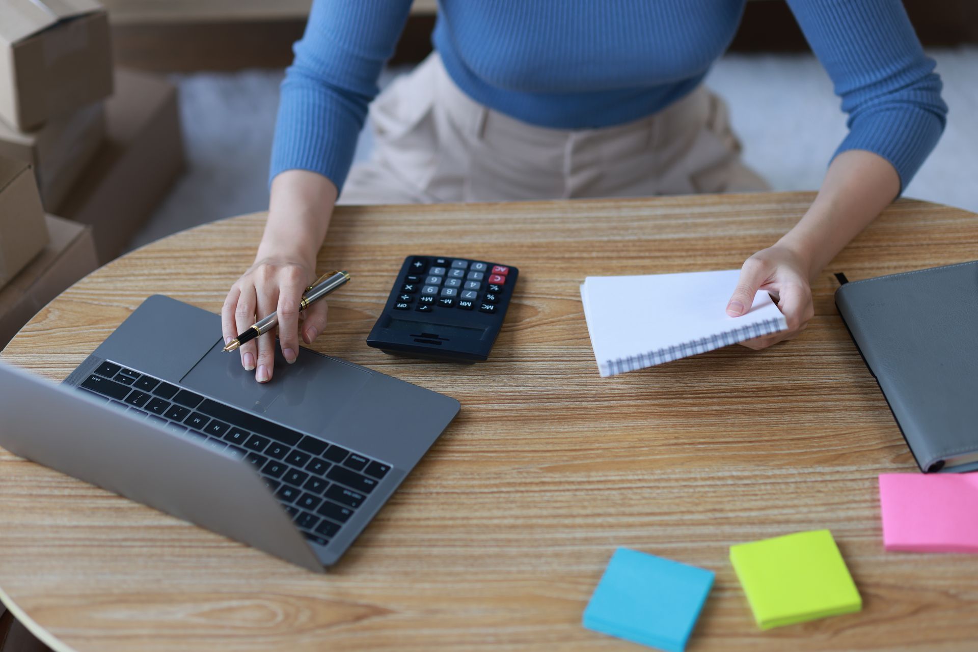 A woman is sitting at a table with a laptop , calculator , notebook and pen.