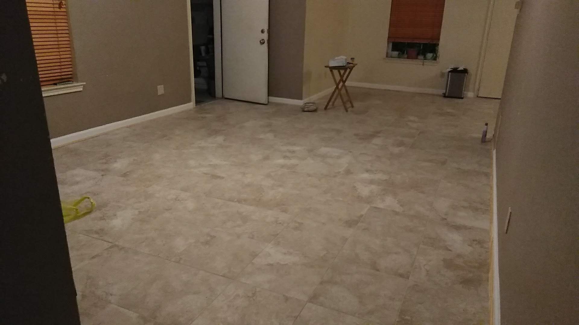 Empty room with beige floor and walls; doorway, window with blinds, small table.