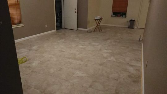 Empty room with beige floor and walls; doorway, window with blinds, small table.
