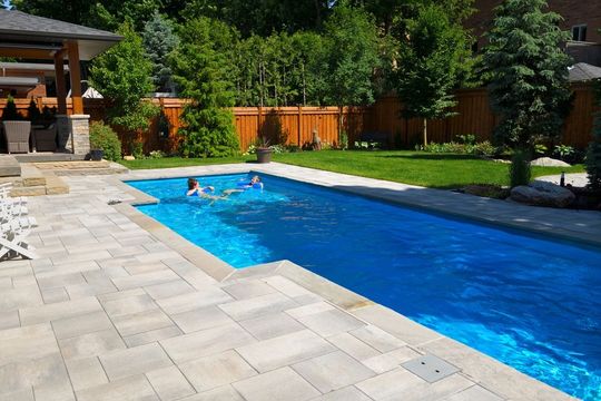 A rectangular swimming pool with two people swimming in it, surrounded by a paved patio and green lawn.