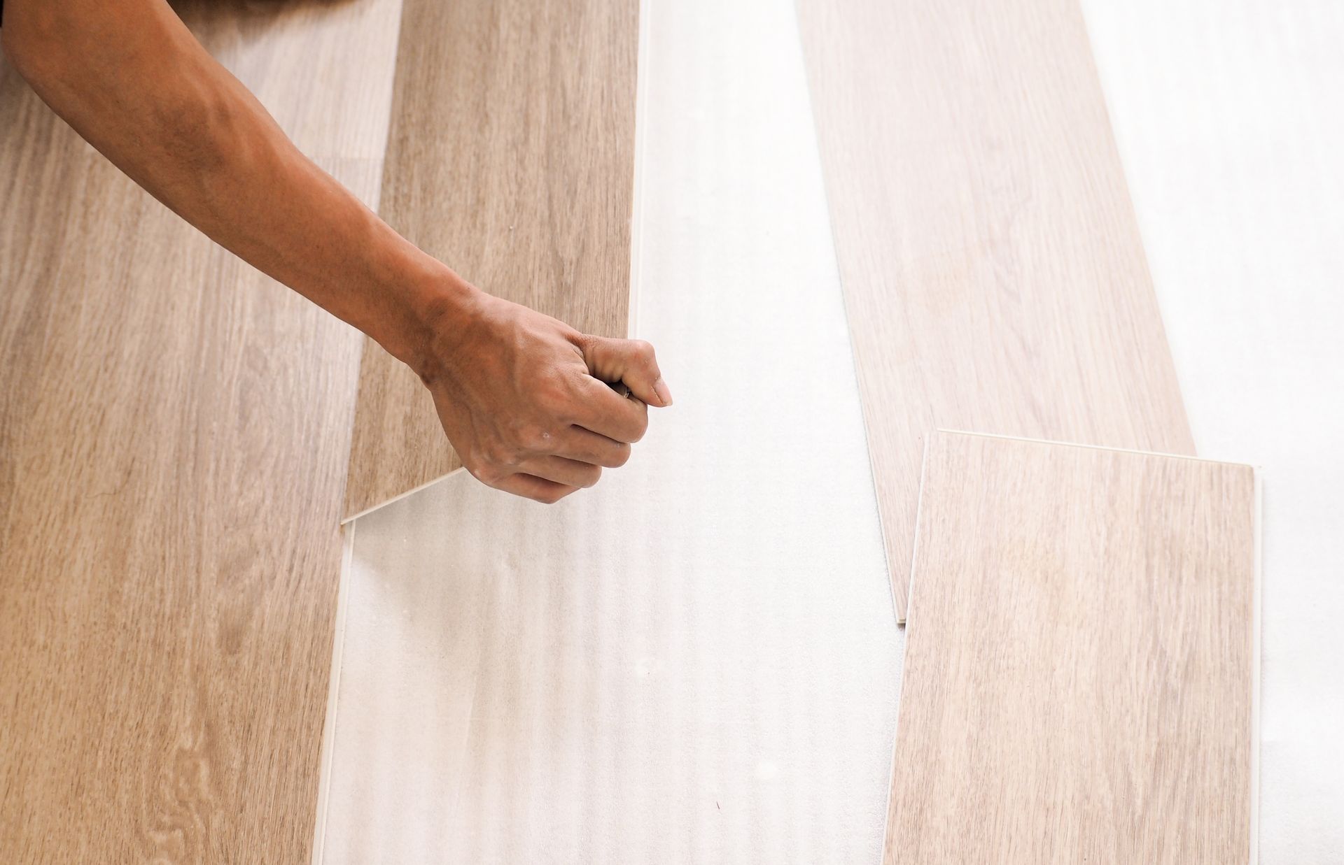 Person installing light-colored wood flooring; hands holding plank against wall.