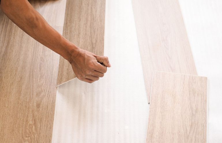 Person installing light-colored wood flooring; hands holding plank against wall.