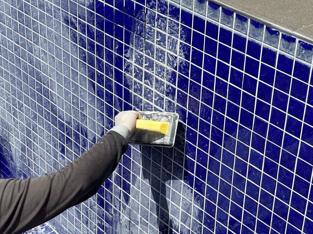 Person grouts blue tile on a pool wall using a yellow float, white grout lines.