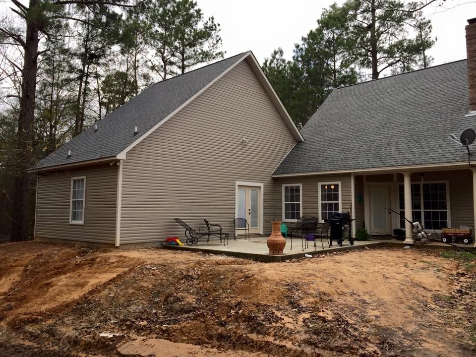 Beige house with gray roof, patio, and surrounding bare earth. Trees in the background.