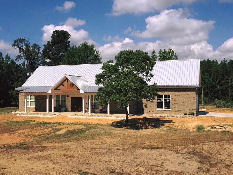 Brick ranch house with a metal roof and a porch, set on a barren landscape, under a partly cloudy sky.