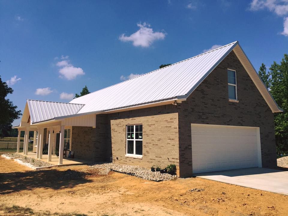New construction brick house with white metal roof, porch, and garage door. Blue sky with clouds.