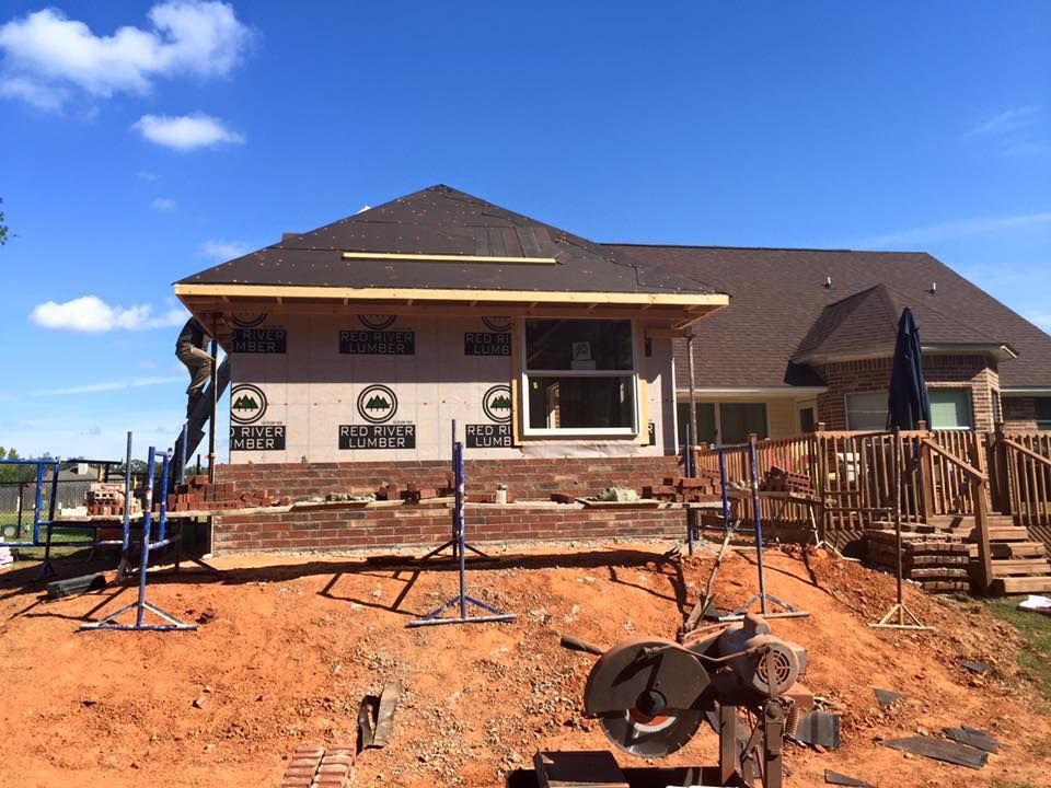 Construction of a brick-faced addition to a house; workers on scaffolding, red clay ground, blue sky.