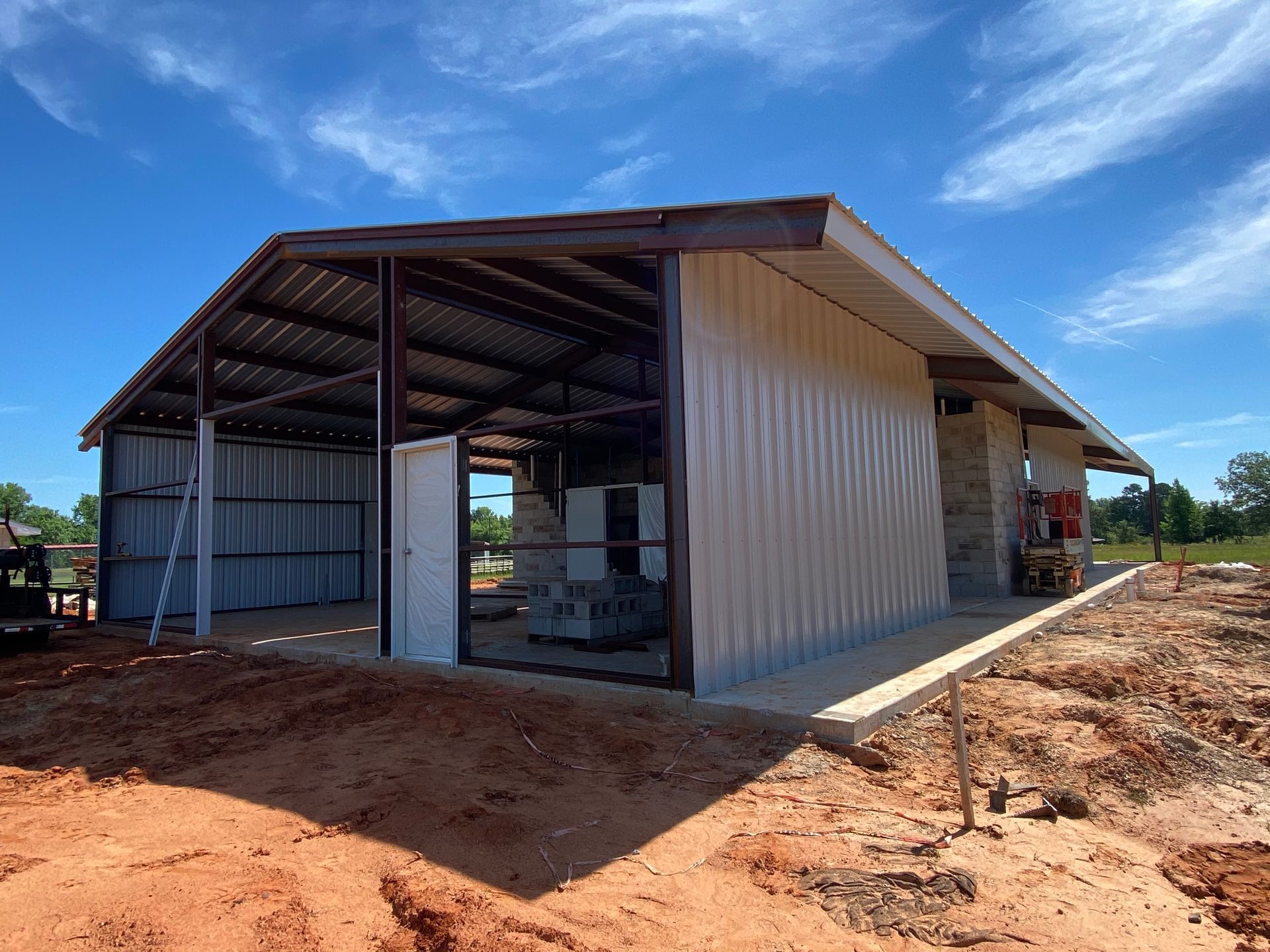 Metal shed under construction on a sunny day. Red dirt, blue sky, brown roof.