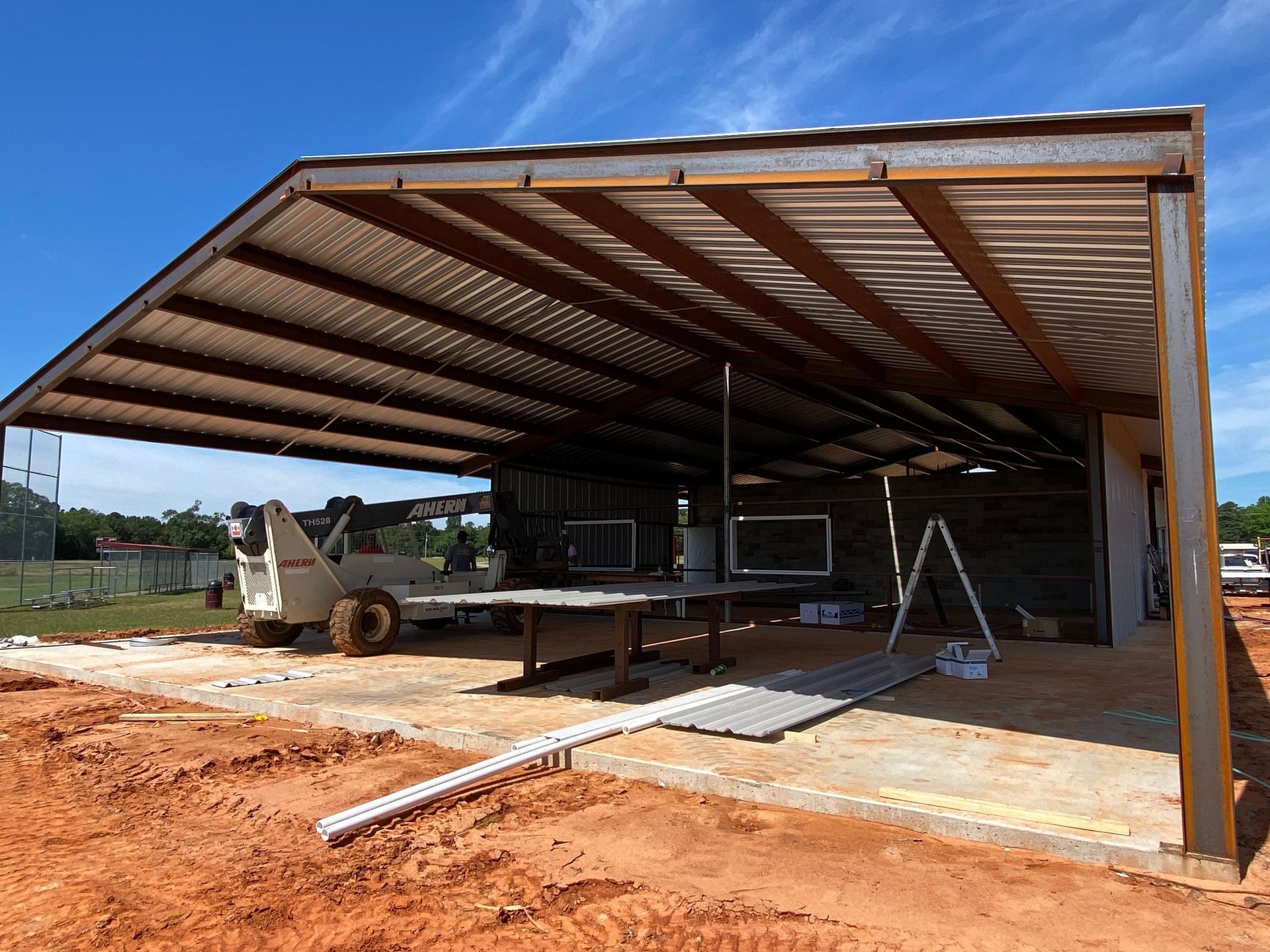 Construction of an open-air metal shed with a red dirt ground under a blue sky.