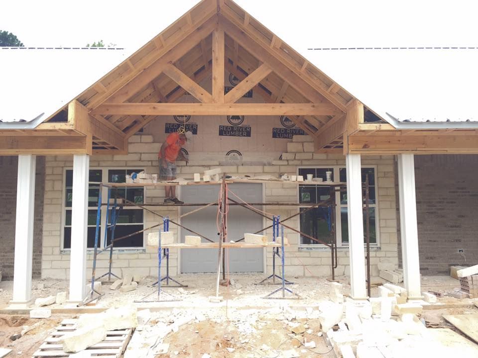 Construction worker on scaffolding laying blocks above a doorway, with completed stone columns and roof.
