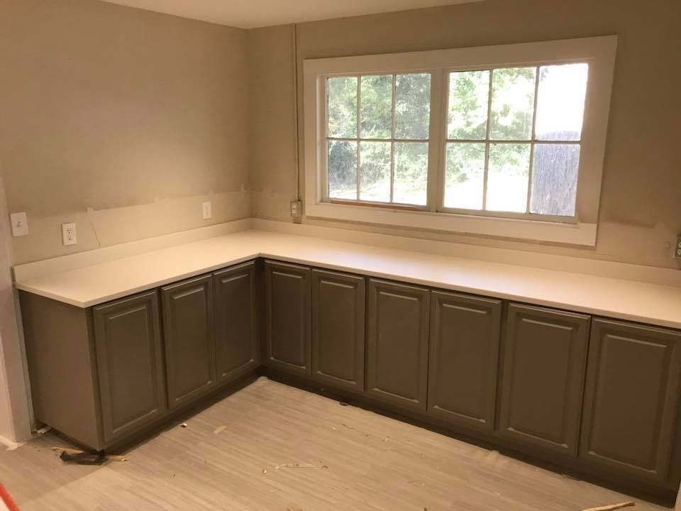 L-shaped, gray kitchen cabinets with light countertops under a window in a room with light flooring and tan walls.