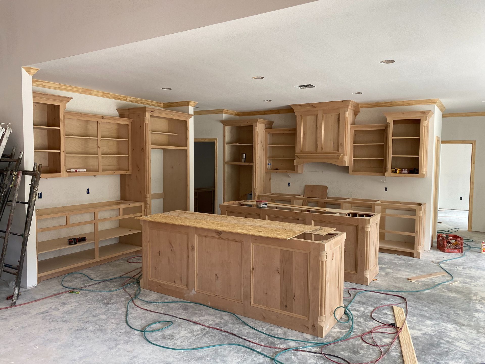 Unfinished kitchen with light wood cabinets and island, under construction, exposed wires, and concrete floor.