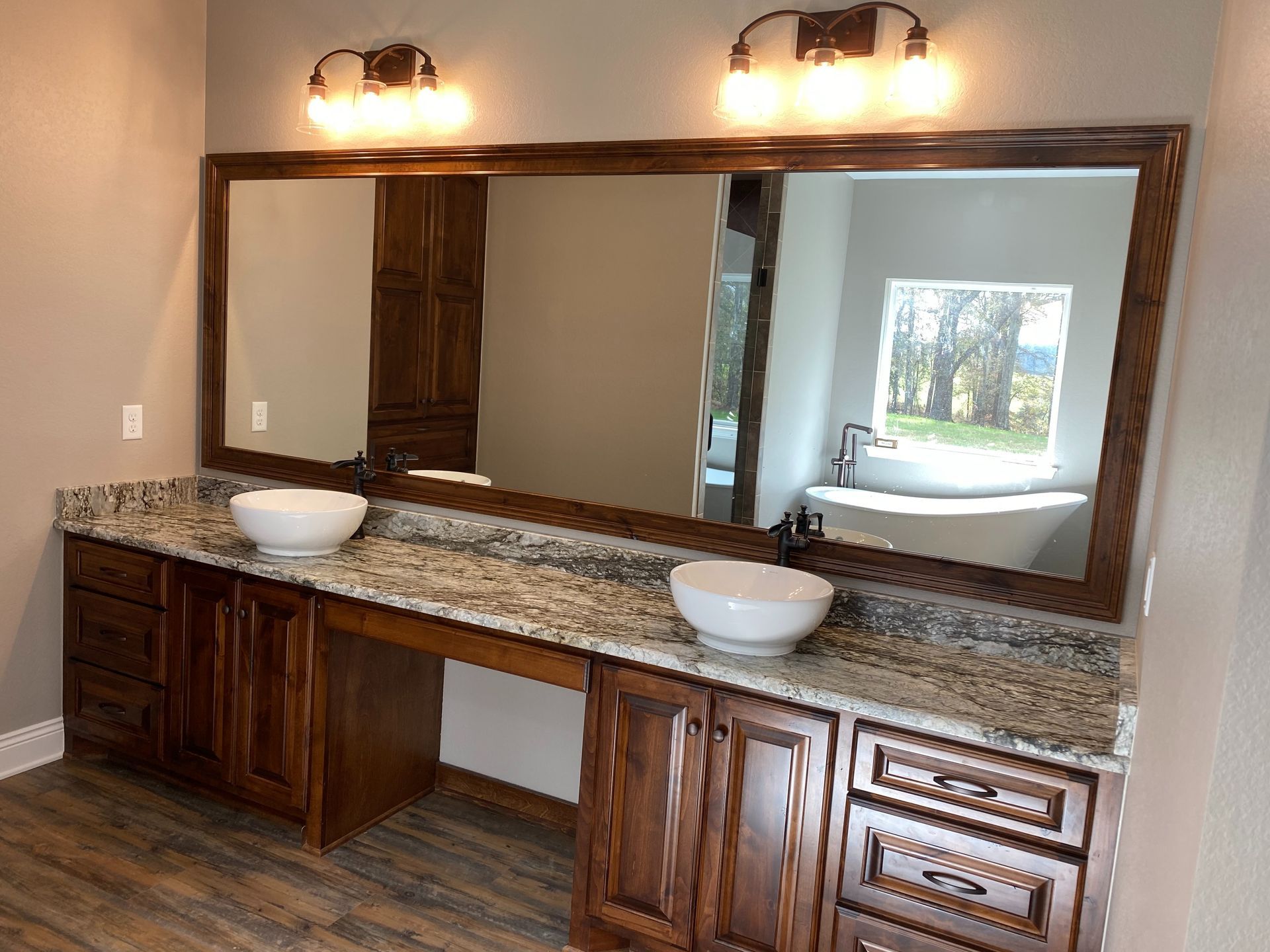 Bathroom vanity with brown cabinets, granite countertop, two vessel sinks, large mirror, and lights.