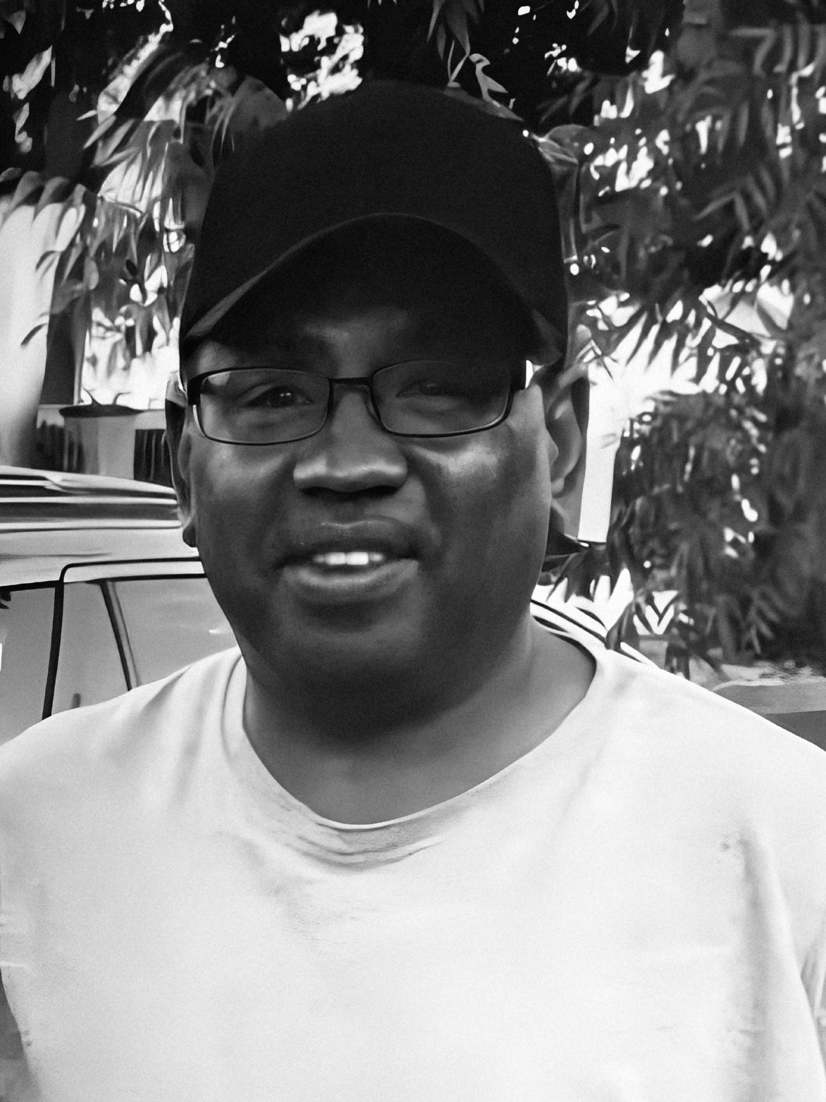 Man wearing glasses and a black cap smiling, standing outdoors near a vehicle.