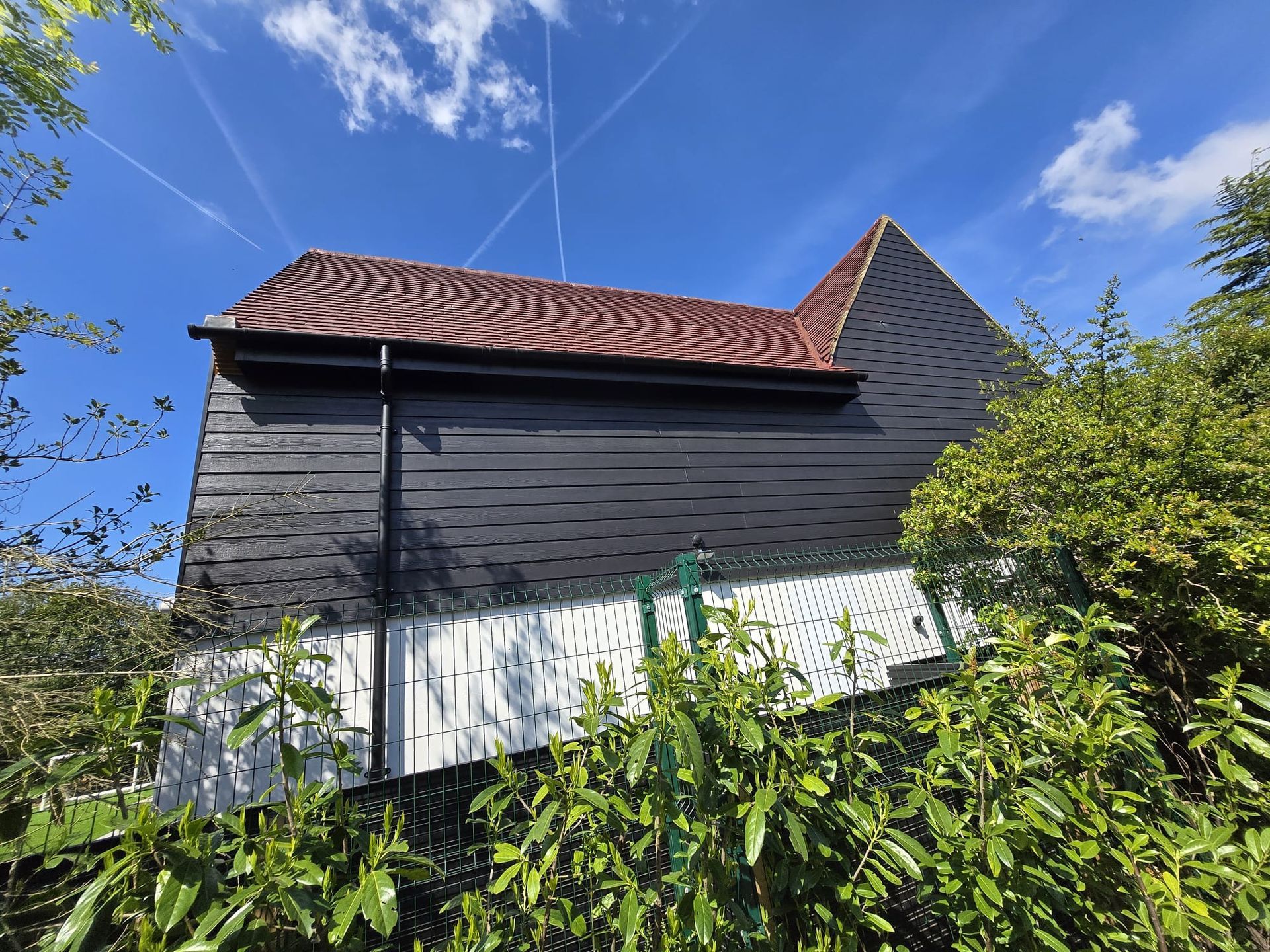 A rustic barn with black horizontal siding and a red tiled roof, framed by lush green foliage against a sunny blue sky.
