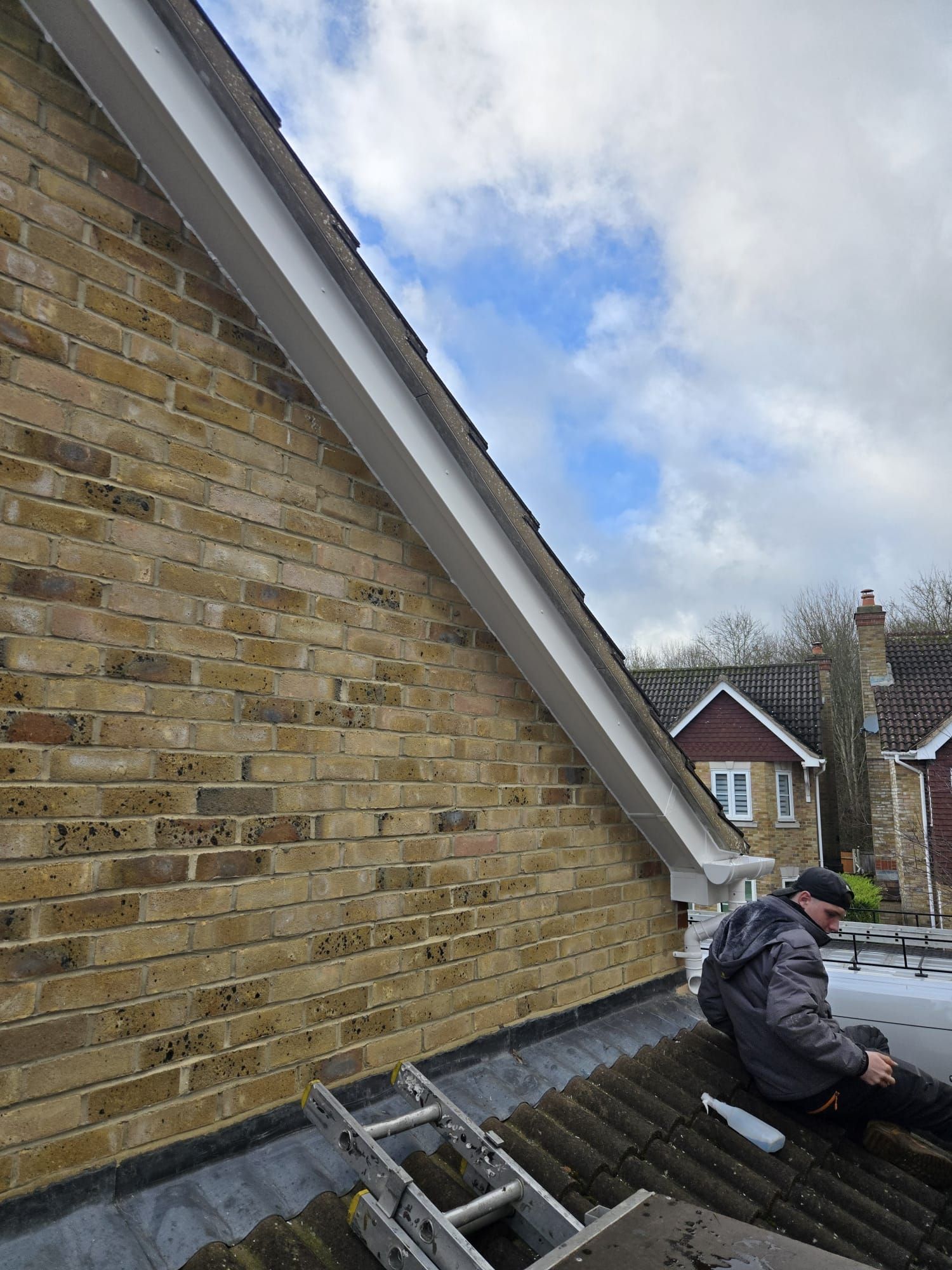 A person sitting on a sloped, tiled roof next to a brick wall and a white bargeboard, overlooking residential houses.