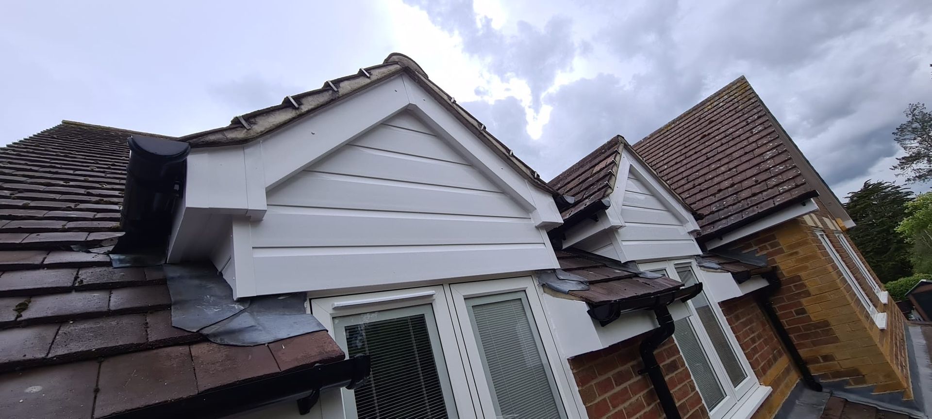 Two white-painted gable dormer windows on a pitched tiled roof, showing exterior brickwork and grey cloudy sky.