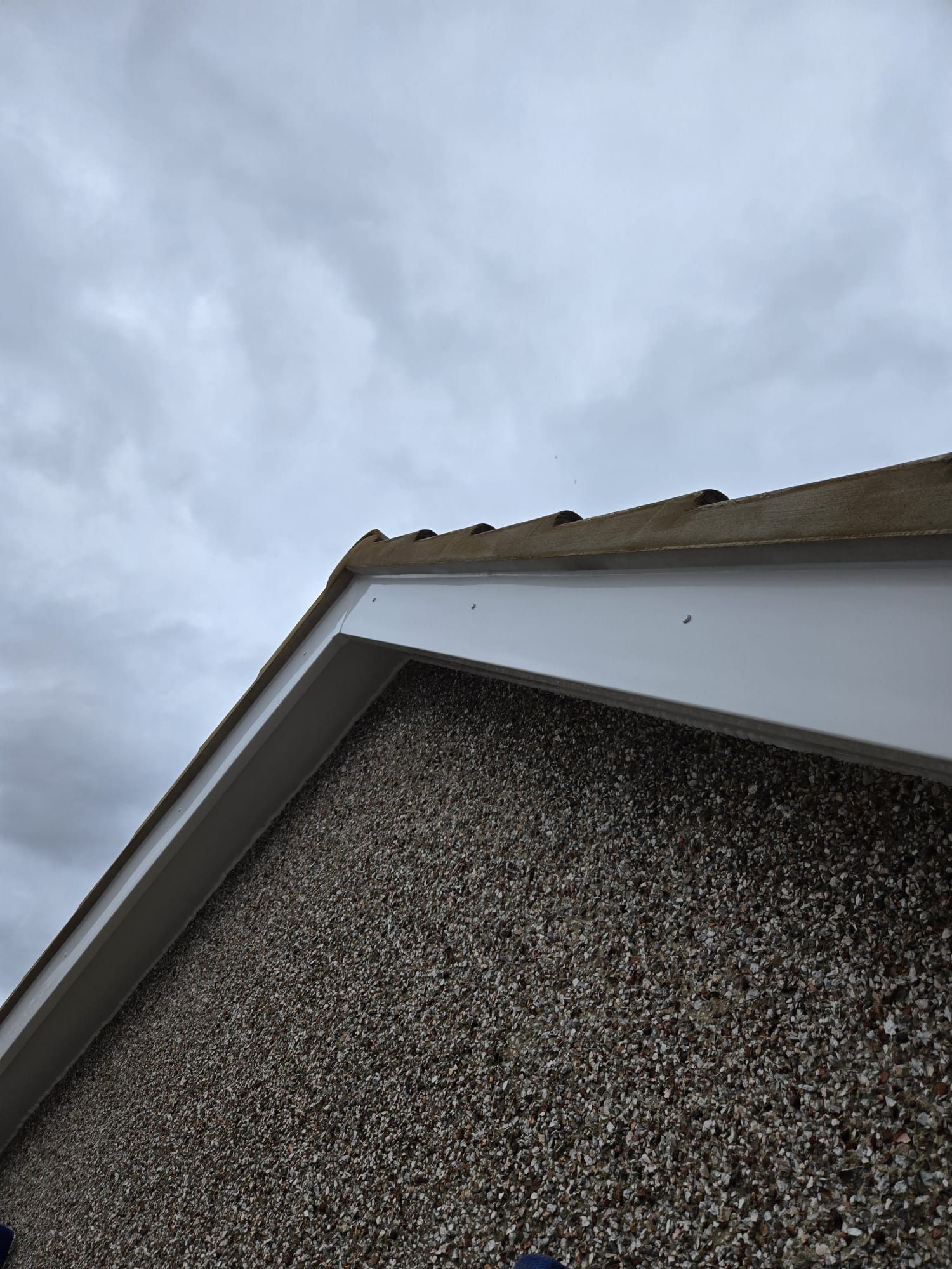 A low-angle view of a pebble-dashed wall meeting a white bargeboard and brown roof tiles under a cloudy sky.