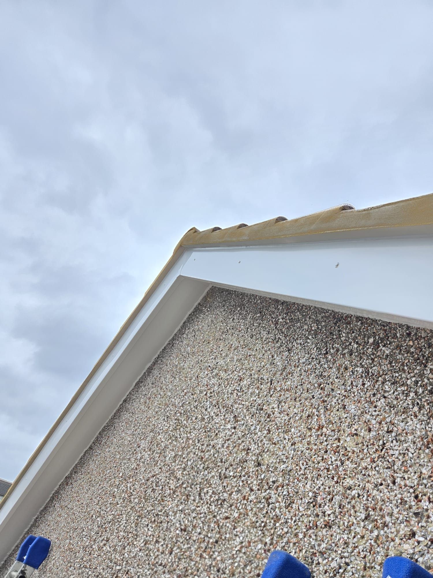 Low-angle view of a pebble-dash house wall meeting a white bargeboard and a brown tiled roof under a cloudy sky.