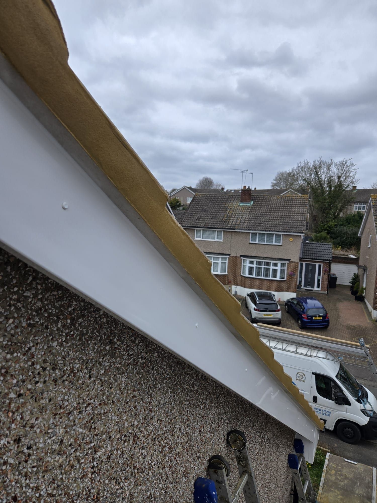 A high-angle view of a white bargeboard and brown roof tiles on a house, looking down at a suburban street with parked cars.