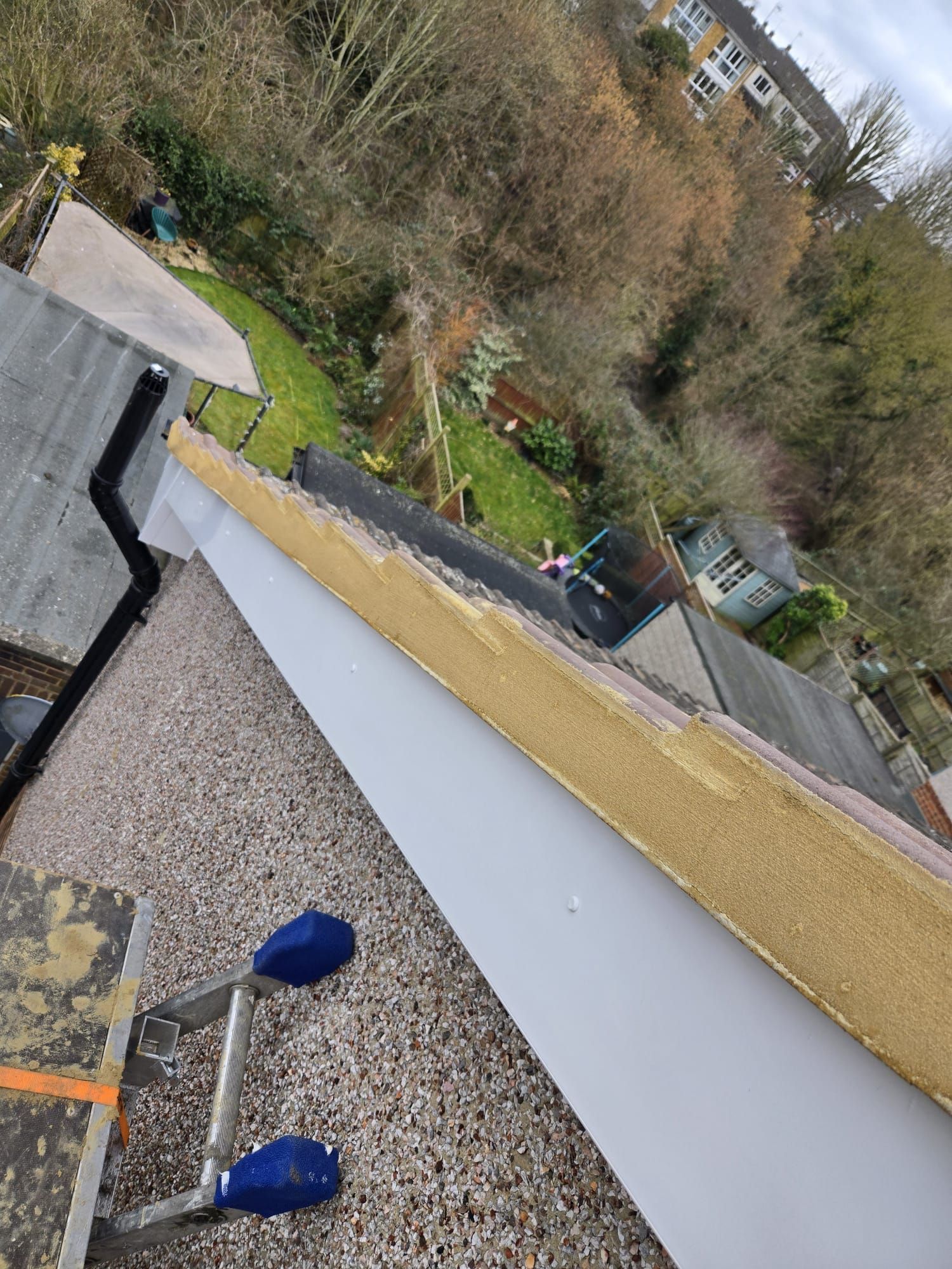 A high-angle view of a roof edge under repair, showing exposed wood fascia and a white soffit above a gravel-covered flat roof.