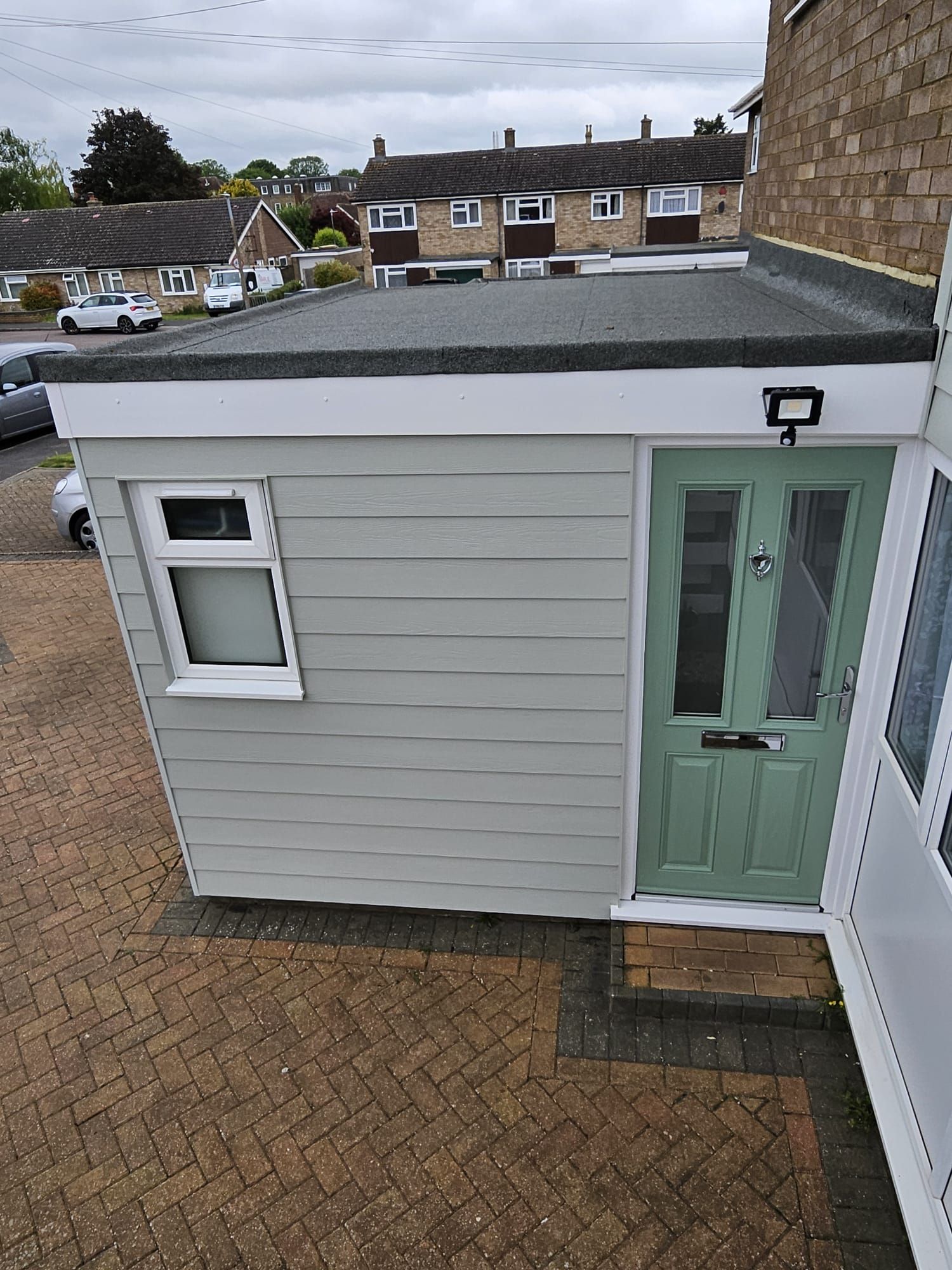 A side view of a residential extension with light gray horizontal siding, a light green front door, and a flat roof.