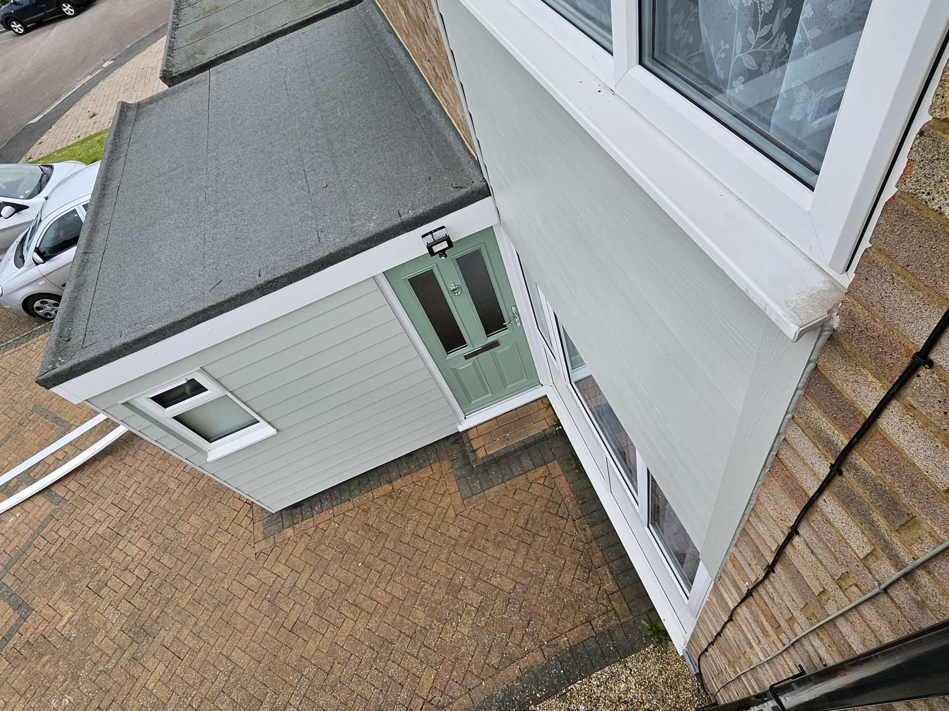A high-angle view of a sage green home extension with a flat roof, a light green door, and paved patio area.