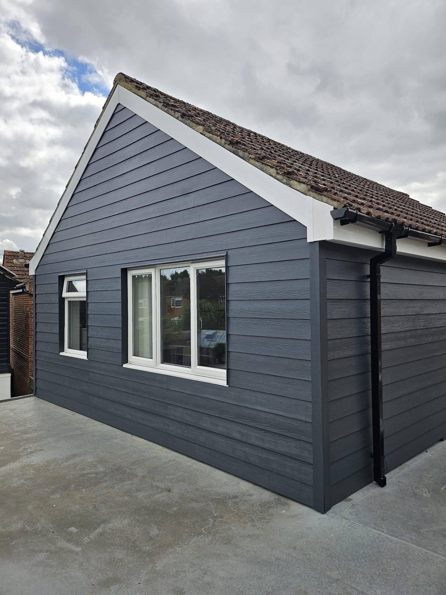 Exterior view of a small building with dark grey horizontal siding, a tiled roof, and white-framed windows.