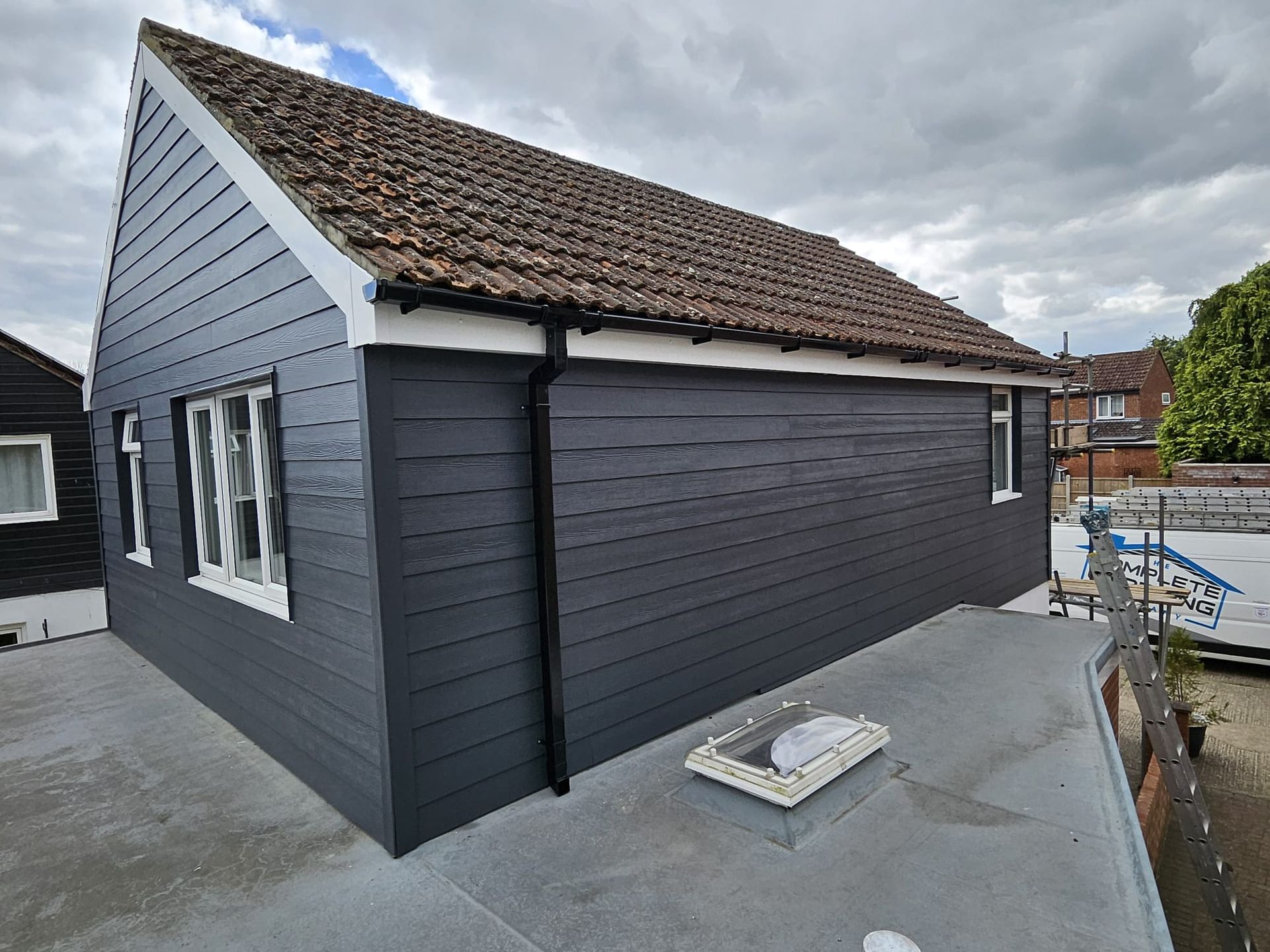 Dark gray building with white trim, brown roof, and skylight on a flat roof under a cloudy sky.