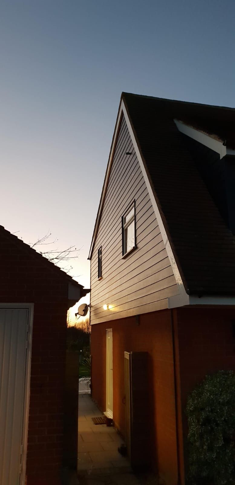 A house with white siding and dark roof, seen from a narrow alley in the evening.