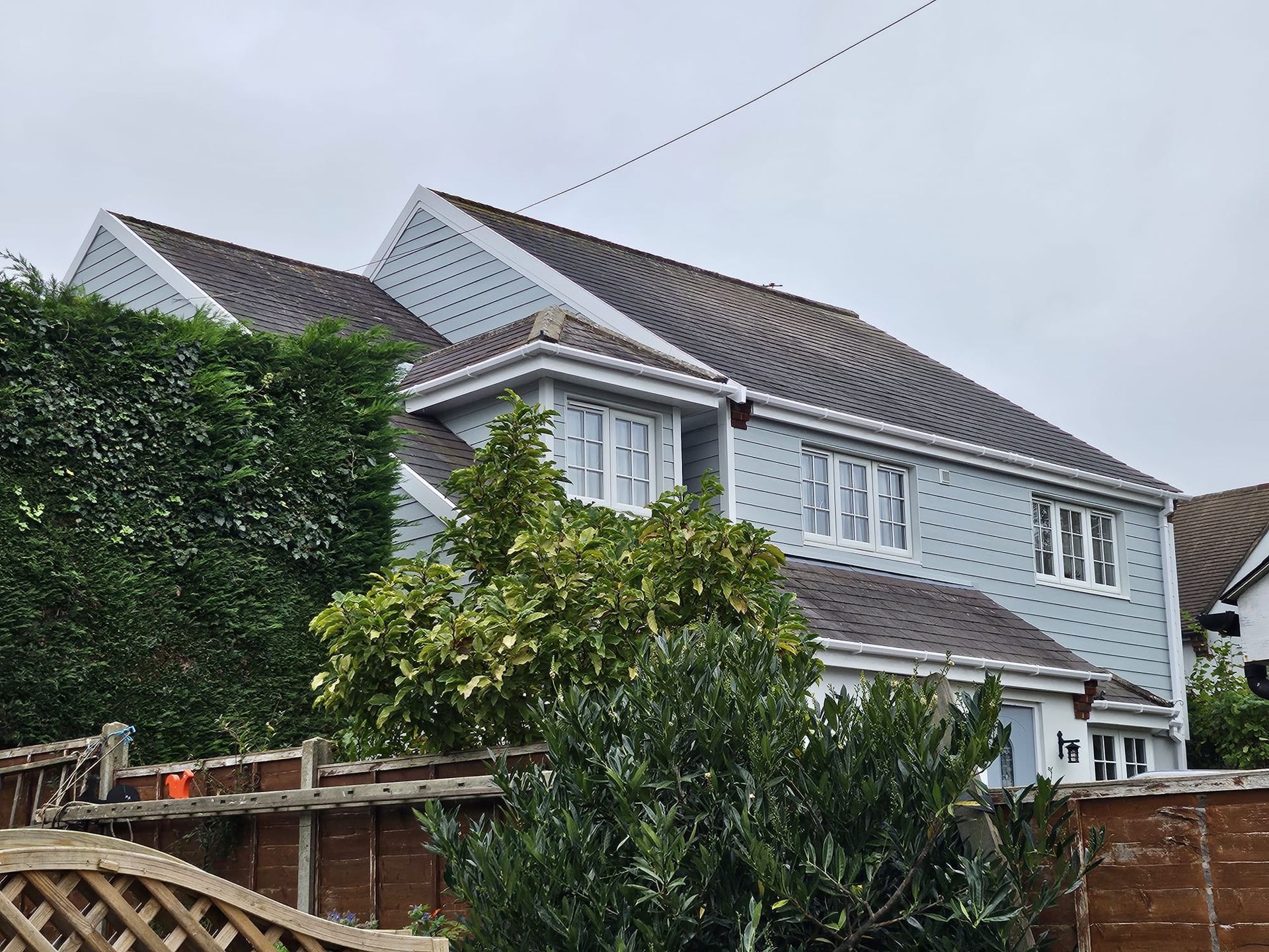 Two-story house with blue siding, gray roof, white trim, and a partial view of hedges and a wooden fence.