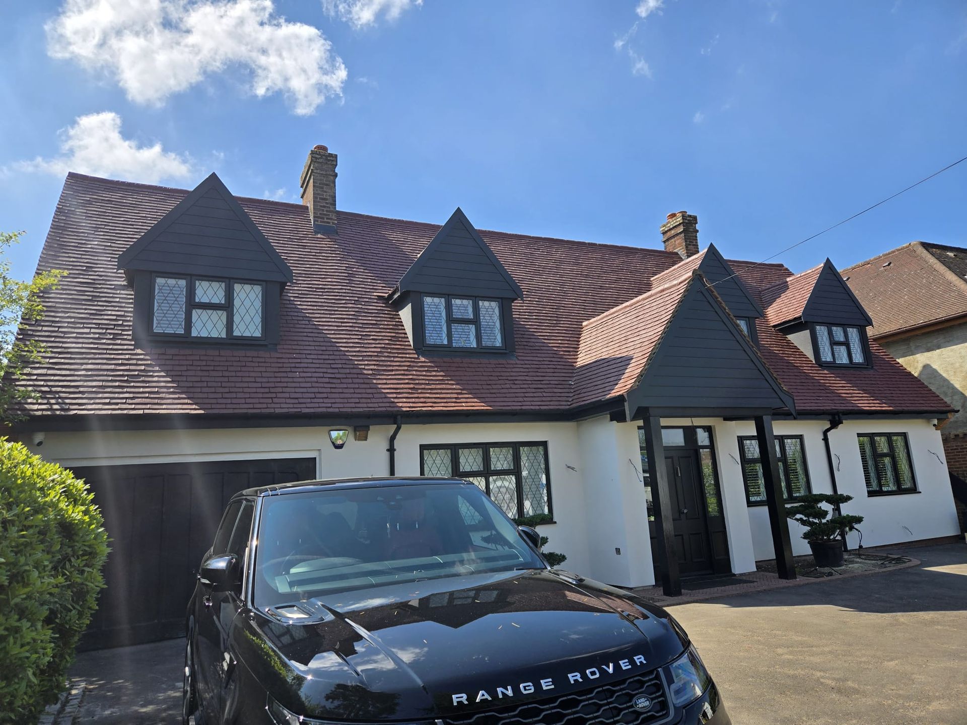 A black Range Rover parked in front of a white two-story house with a red tiled roof, multiple gables, and dormer windows.