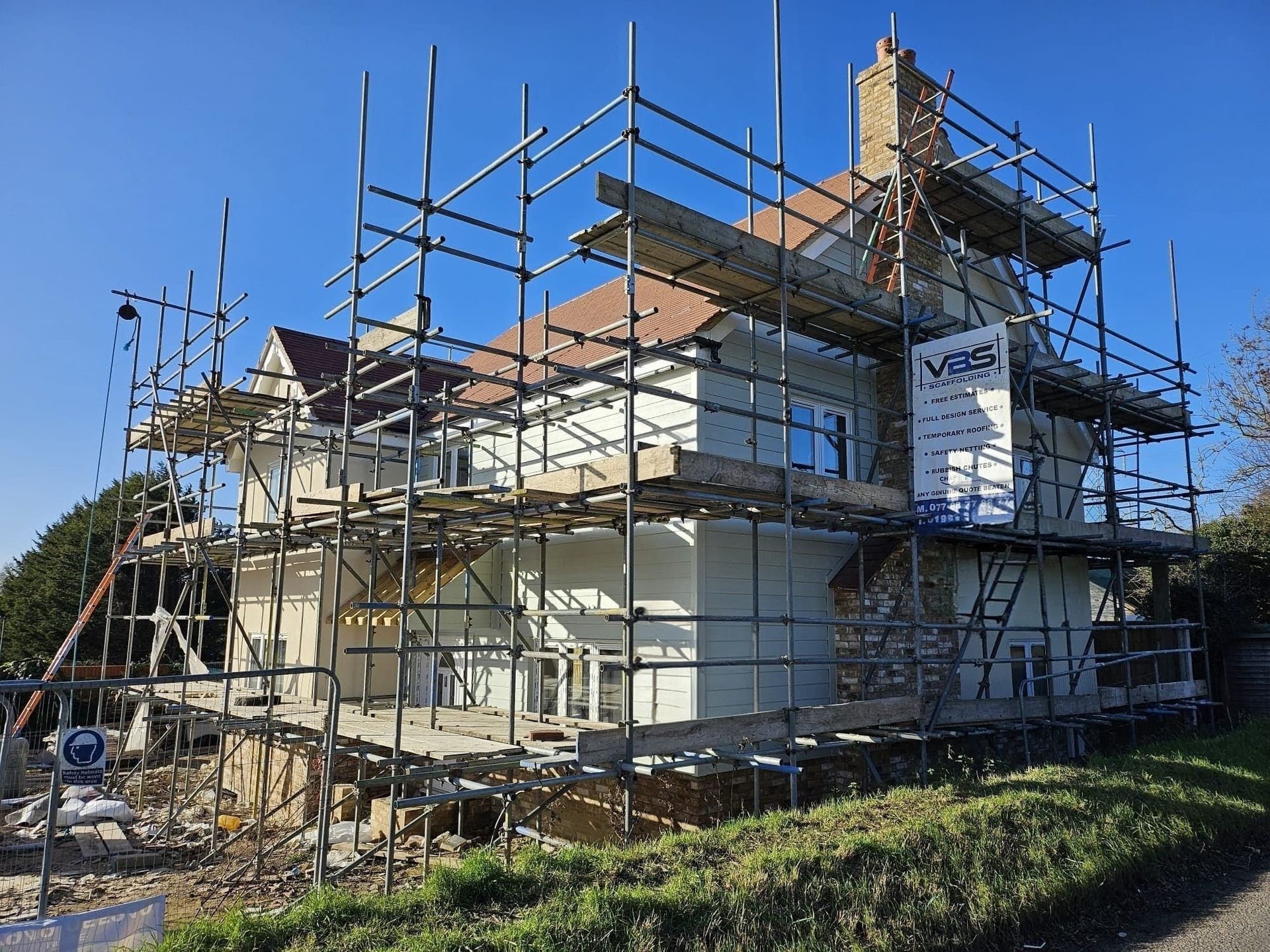 A two-story light-colored house under renovation, surrounded by extensive metal scaffolding under a clear blue sky.