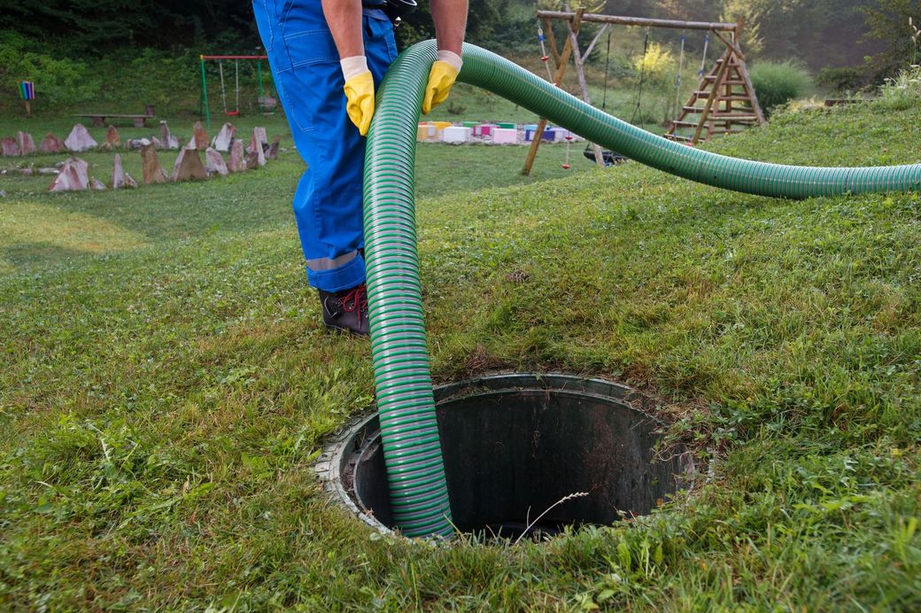 Un operaio con pantaloni blu e guanti gialli usa un tubo per pulire una buca nel prato del cortile.