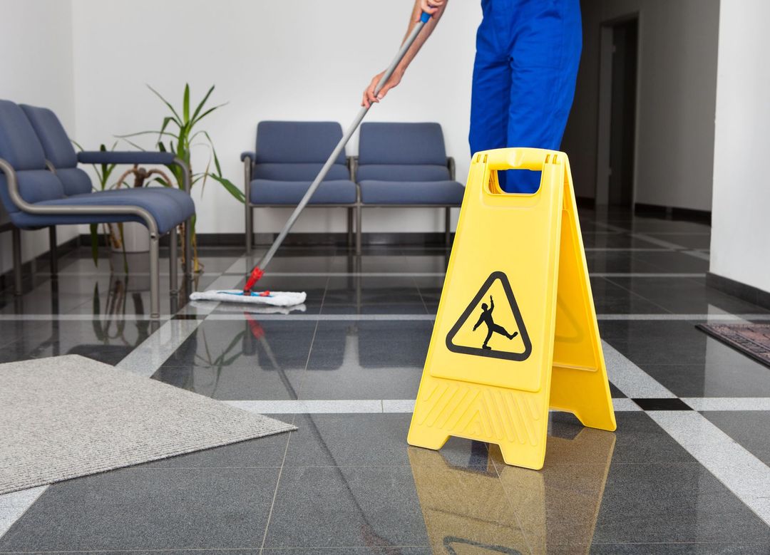 Un operaio lava il pavimento lucido della hall accanto a un cartello giallo di avvertimento per pavimenti bagnati.