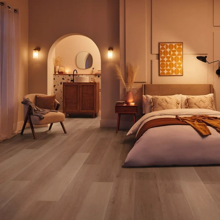 Bedroom with wood-look flooring, bed, chair, and a bathroom visible through an archway. Warm, neutral tones — Mareeba Floor Covering Centre in Cooktown, QLD