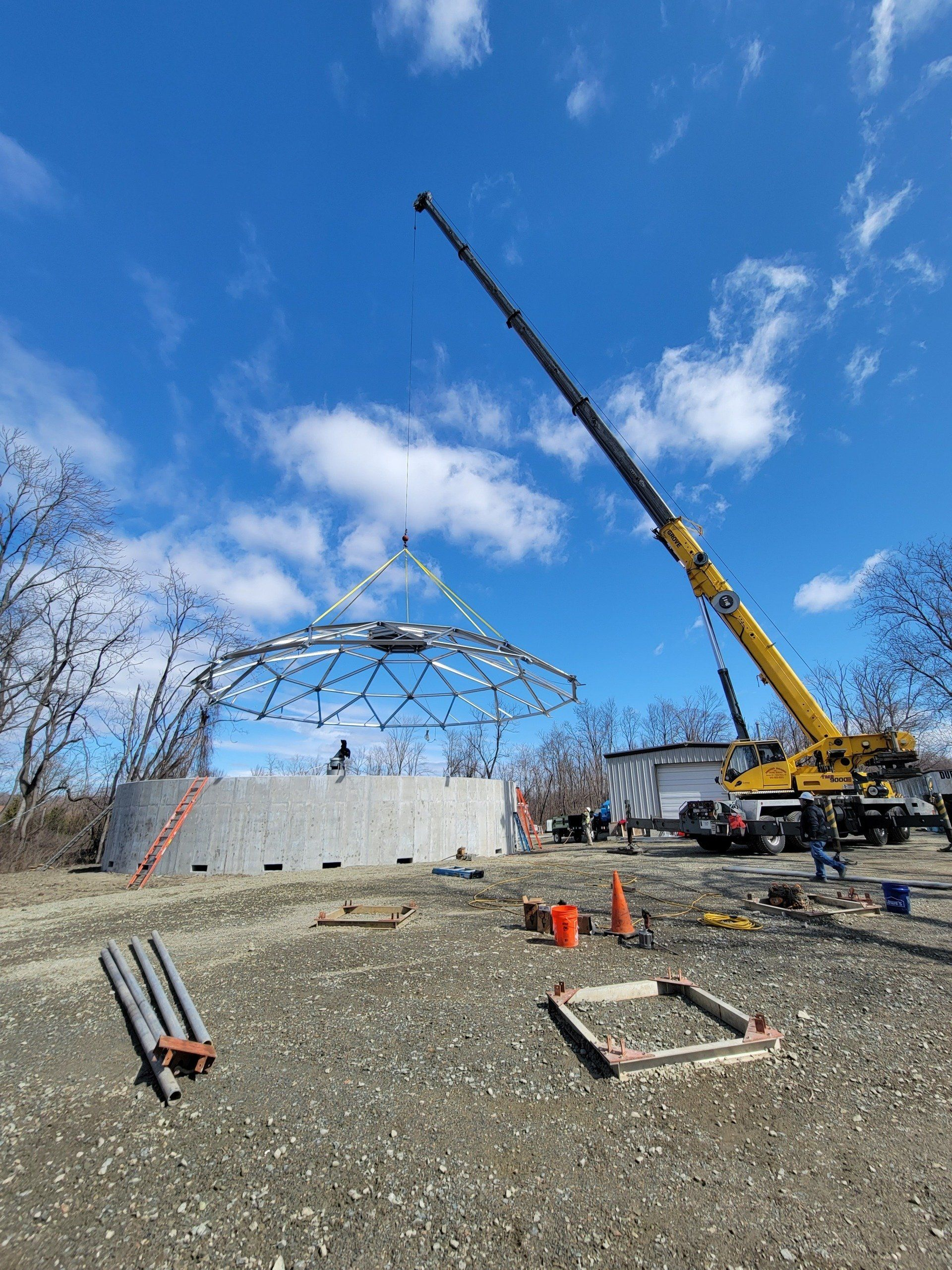 A large yellow crane is lifting a metal structure in a dirt field.