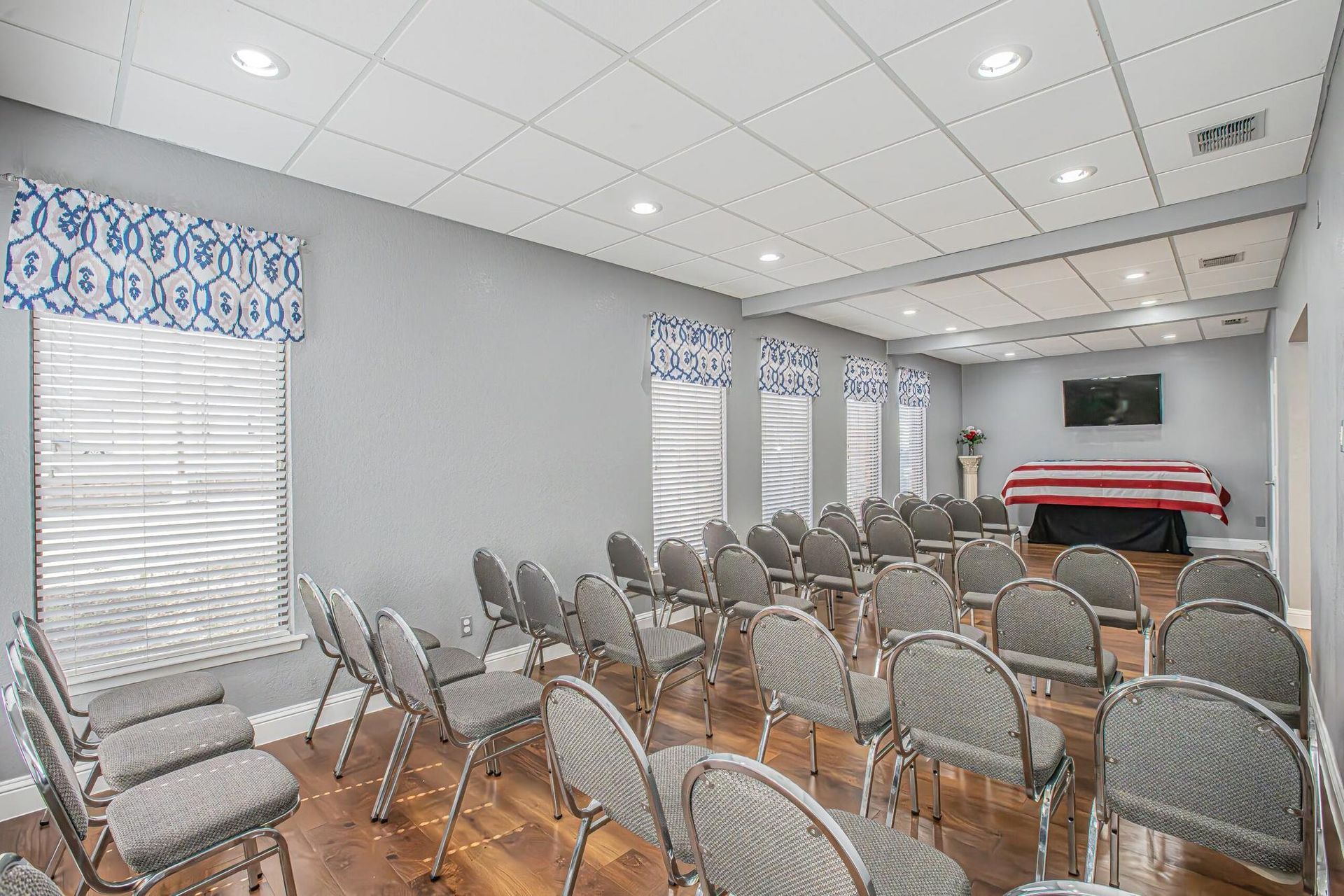 Funeral home chapel with rows of chairs facing a casket; light gray walls, wood floor, and blue accents.