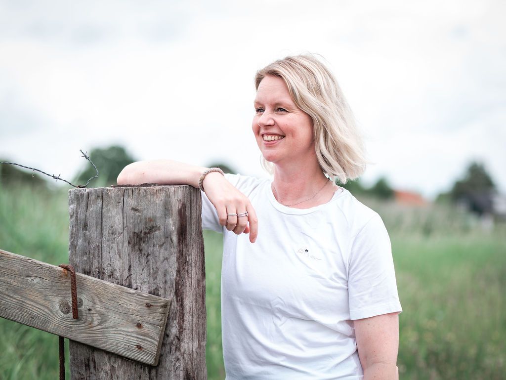 Een blonde vrouw glimlacht en leunt tegen een houten hekpaal in een veld. Yoga Marjolijn Hoogmade