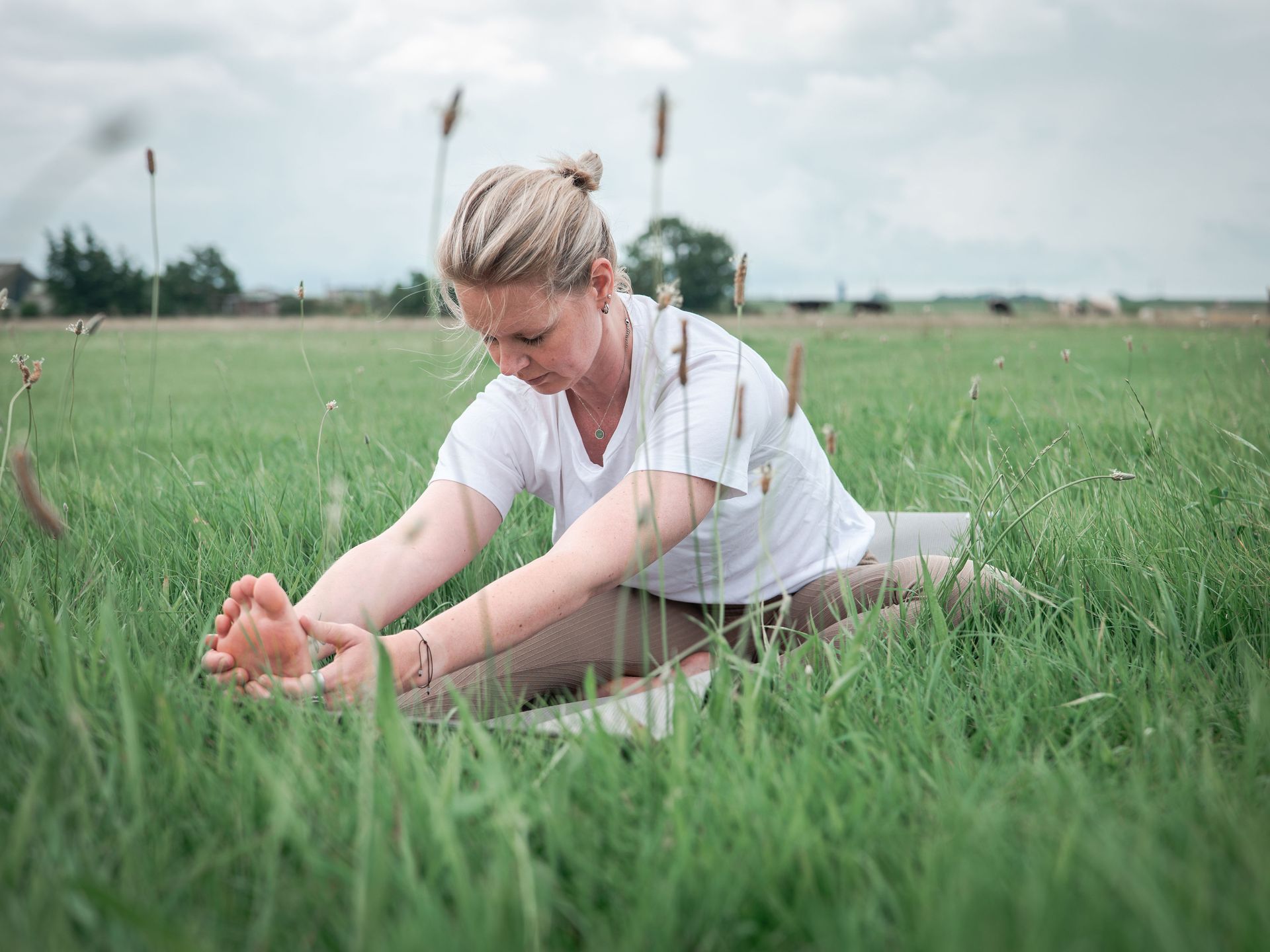 Een vrouw in een wit shirt strekt zich uit in het hoge gras, zit met gespreide benen en reikt naar haar voet. Yoga.