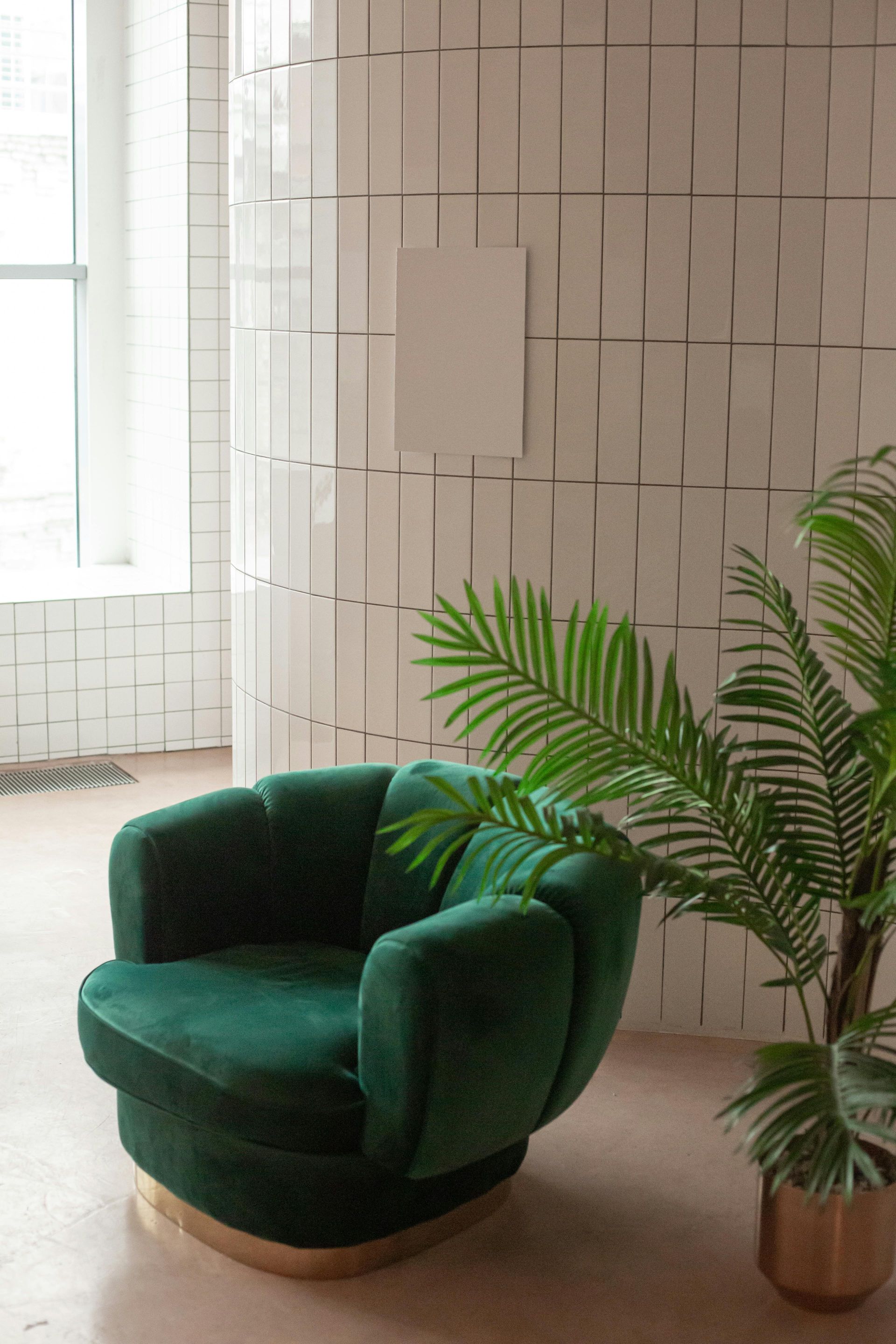 Emerald green velvet armchair, palm plant, and blank white artwork in a modern interior setting.
