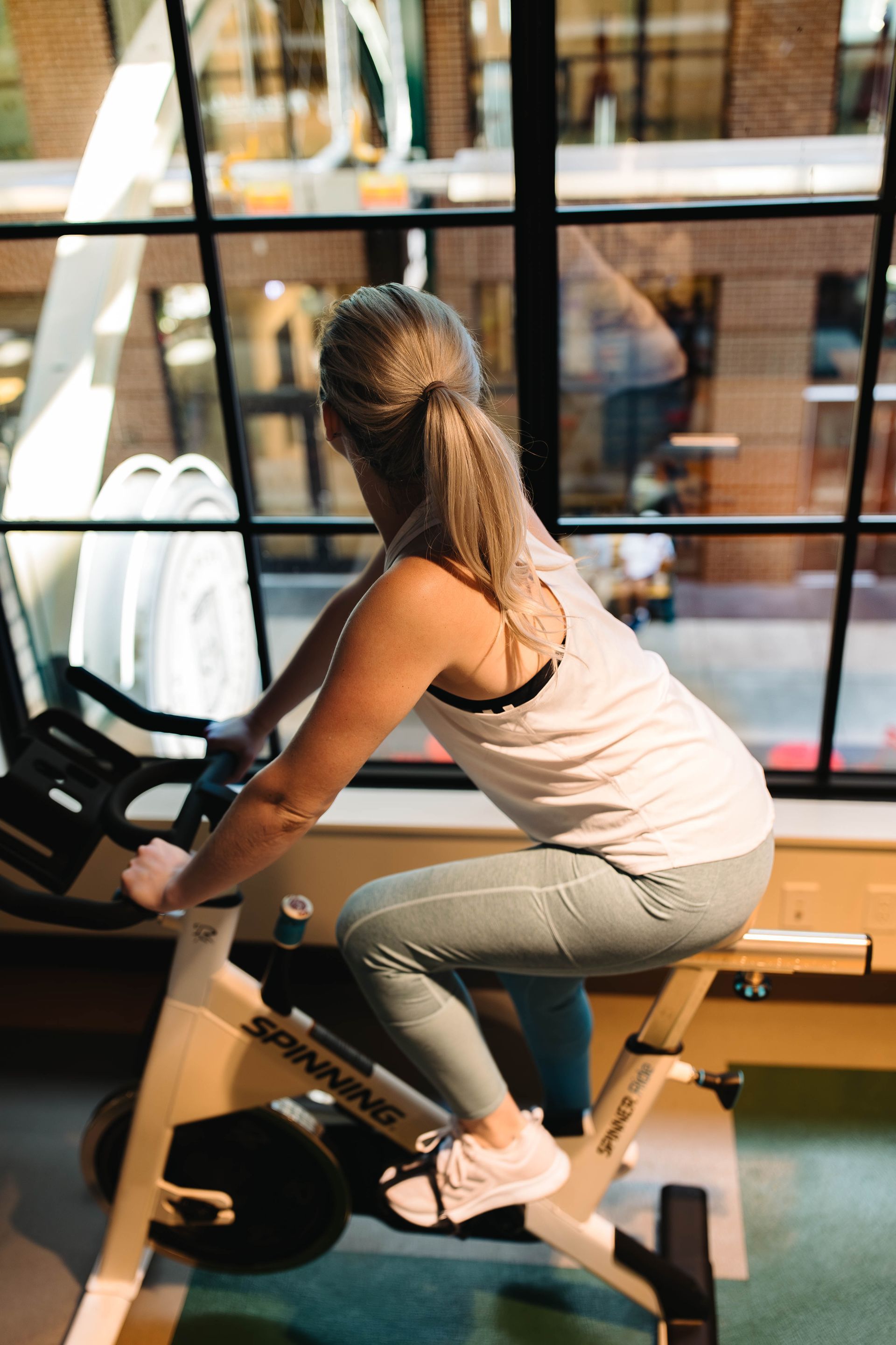 A woman works out on a stationary bike inside the 24-hour fitness studio at Gordon Lofts in Eugene, Oregon. Sunlight streams through the large industrial-style windows, offering a view of the 5th Street Public Market below. The scene blends modern wellness with urban charm, reflecting the active, vibrant lifestyle of Gordon Lofts residents.