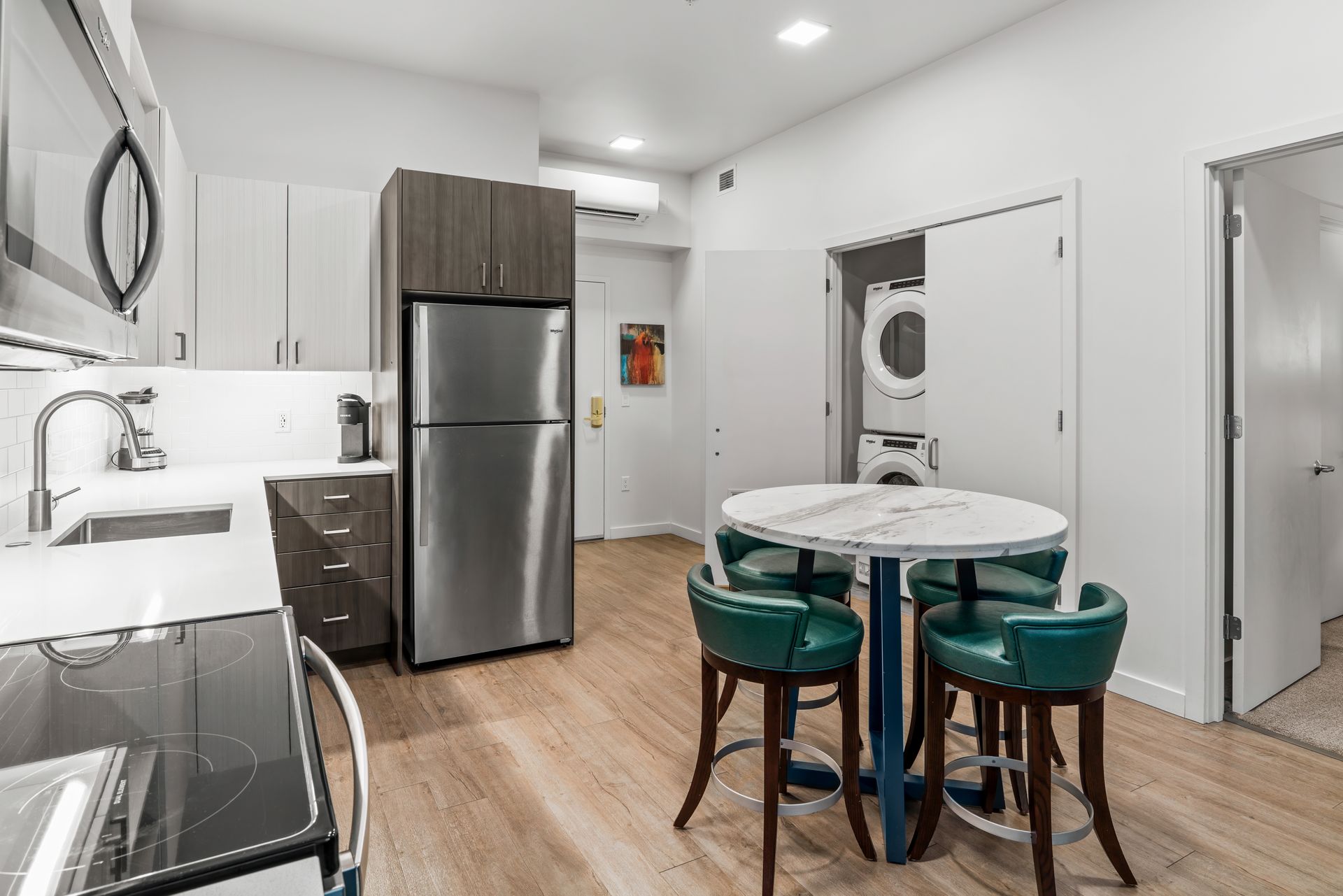 A modern furnished one-bedroom, one-bath apartment at Gordon Lofts in Eugene, Oregon, featuring a sleek kitchen with stainless steel appliances, white quartz countertops, and wood-style flooring. A marble dining table with teal chairs sits beside an in-unit washer and dryer, capturing the effortless convenience and refined comfort of downtown loft living.