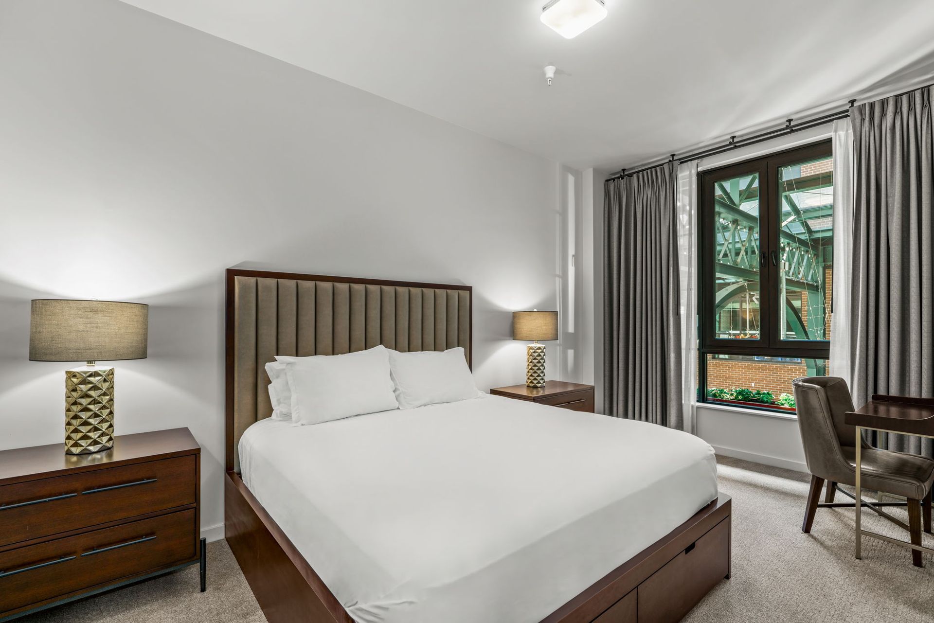 Modern bedroom in a move-in ready apartment at Gordon Lofts in Eugene, OR, featuring a plush upholstered headboard, sleek wood furniture, and soft neutral tones. A large window with gray curtains frames a view of the Market District’s iconic green steel architecture, blending urban vibrancy with cozy comfort.
