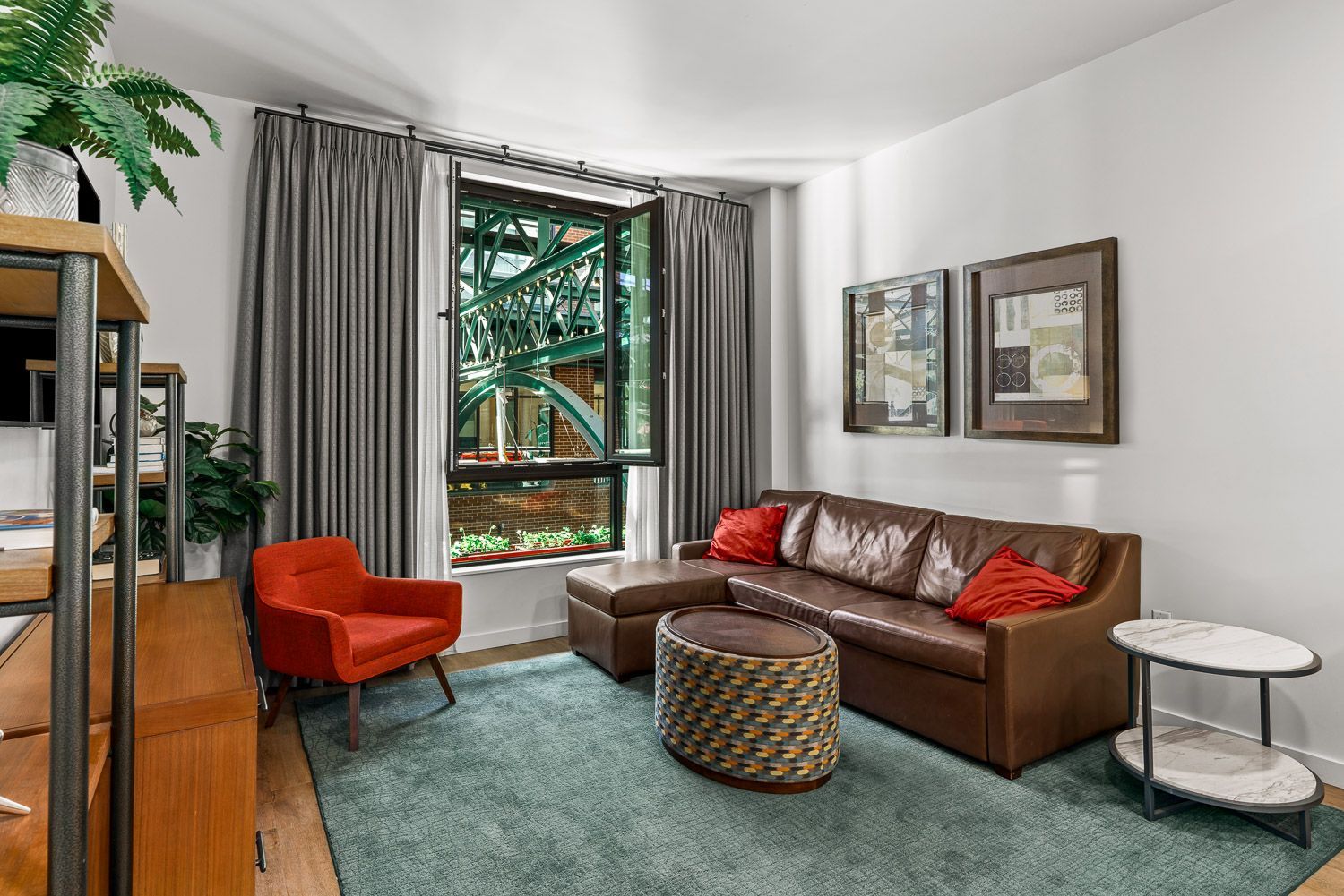 Cozy living room at Gordon Lofts in Eugene, OR, featuring a brown leather sectional with red accent pillows, a woven round coffee table, and an orange mid-century modern chair. A large window with gray curtains offers a unique view of the Market District's architectural details. Decor includes framed wall art, a green area rug, and a sleek two-tiered marble side table, creating a comfortable and stylish atmosphere.