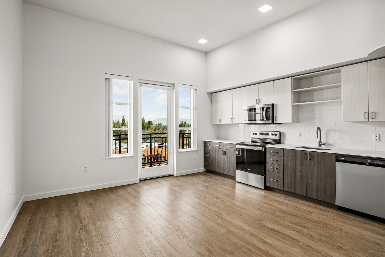 Bright and modern kitchen at Gordon Lofts in Eugene, OR, featuring stainless steel appliances, sleek two-tone cabinetry, and white subway tile backsplash. The space opens to a private balcony through a glass door and large windows, offering scenic views and abundant natural light. Warm wood-style flooring adds to the clean, contemporary aesthetic.
