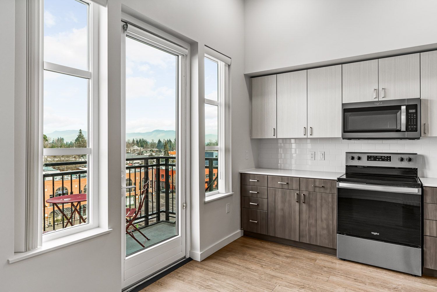 Bright and airy kitchen at Gordon Lofts in Eugene, OR, showcasing modern two-tone cabinetry, stainless steel appliances, and wood-style flooring. A glass door and tall windows open onto a private balcony with bistro seating, offering views of the surrounding Market District and distant hills, creating a seamless indoor-outdoor living experience.