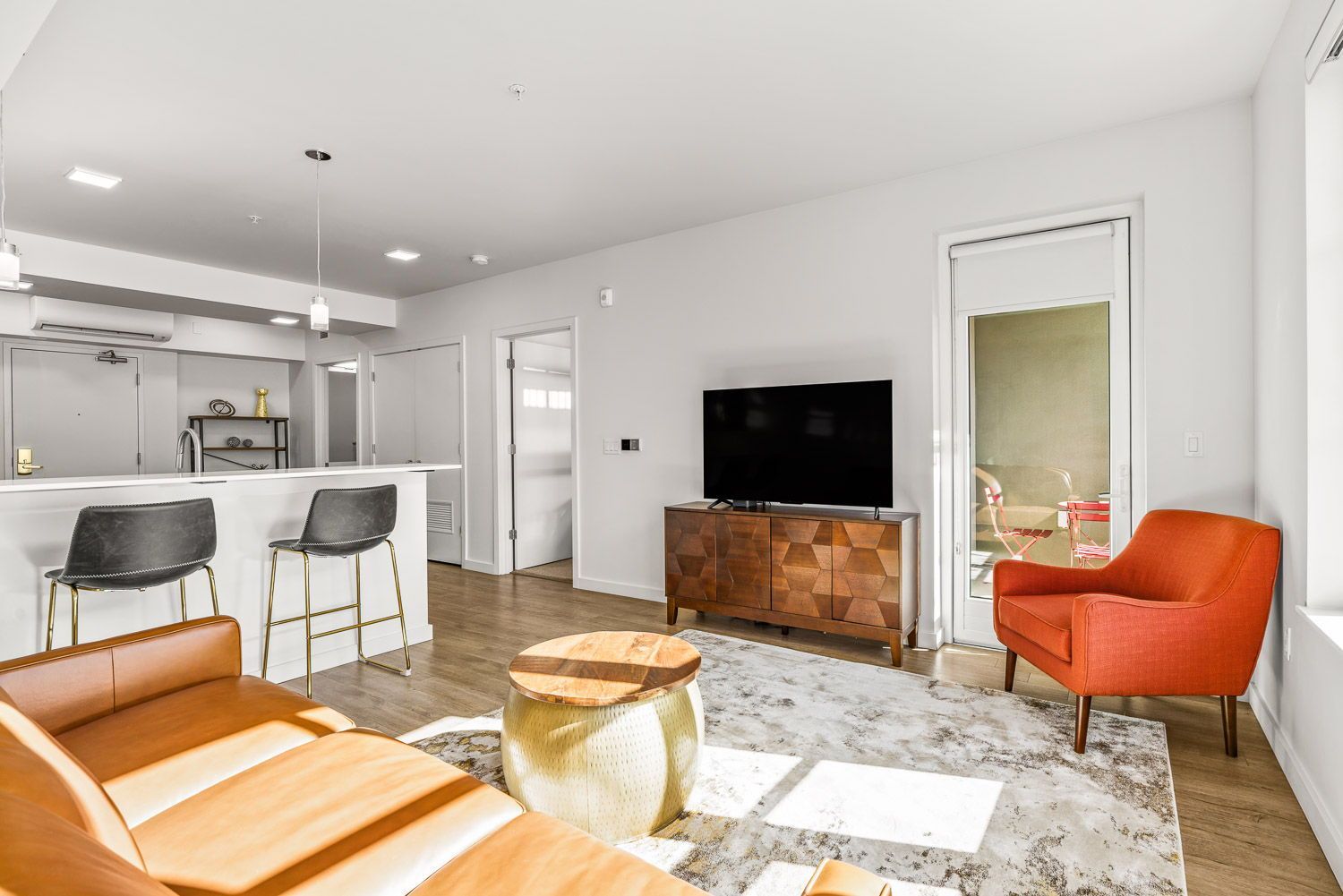 Bright and inviting living space at Gordon Lofts in Eugene, OR, featuring a mid-century modern design with a leather sofa, geometric wood media console, and a bold orange accent chair. The room opens to a private balcony with bistro seating, and includes a cozy breakfast bar with two stools, pendant lighting, and warm wood-style flooring throughout.