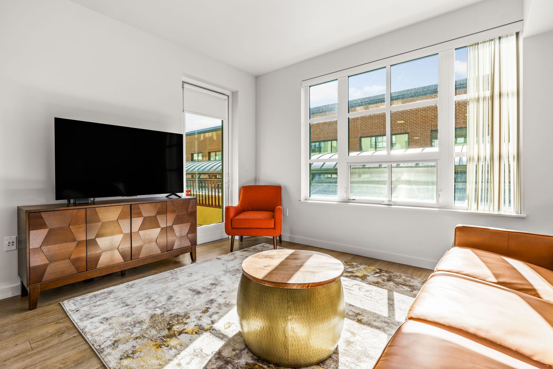 Sunlit living room at Gordon Lofts in Eugene, OR, featuring a mid-century modern aesthetic with a geometric wood console, flat-screen TV, leather sofa, bold orange accent chair, and round gold coffee table. Large windows and a glass door fill the space with natural light and offer views of the surrounding 5th Street Public Market area.