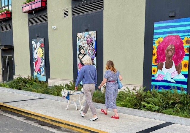 A couple walks a white husky past a colorful outdoor art gallery on the side of the Gordon Lofts building in Eugene, Oregon. The sidewalk is lined with lush ferns, while vibrant murals—featuring butterflies, bold portraits, and pop-art designs—add creative flair to the modern facade. The scene captures the neighborhood’s artistic spirit and walkable charm.